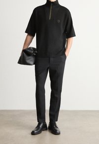 Černý krátký rukáv top s vysokým límcem a zlatým zipem; spárován s černými tailored kalhotami a černými šněrovacími botami. Drží černou tašku.