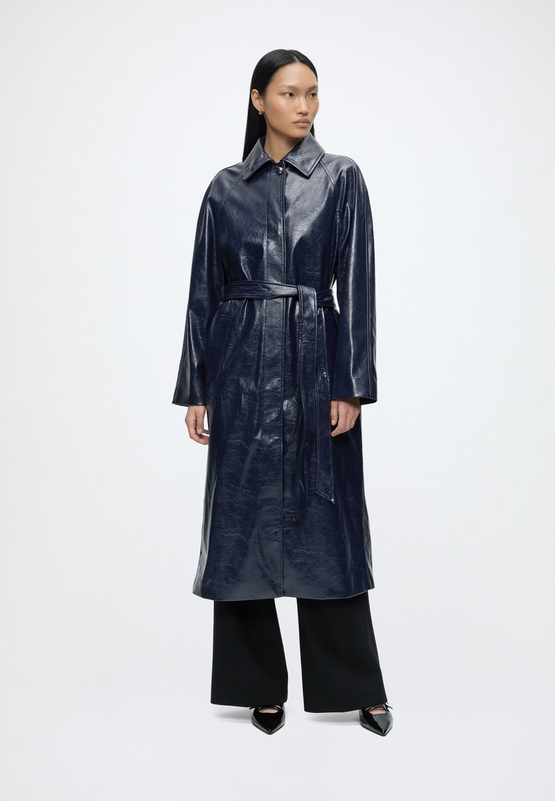 Femme portant un long manteau en cuir bleu marine ceinturé, un pantalon noir à jambes larges et des chaussures noires pointues, debout devant un fond blanc uni.