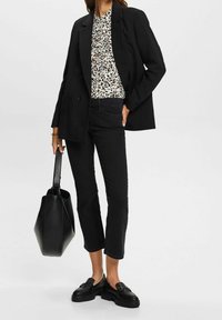 Mujer con blazer negro, blusa con estampado de leopardo, jeans negros recortados, mocasines negros, sosteniendo un gran bolso de mano negro de cuero.