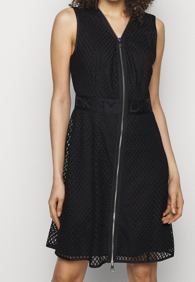 Robe noire sans manches avec fermeture éclair à l'avant, présentant un motif texturé en losanges et une bande à logo DKNY à la taille. Silhouette trapèze.