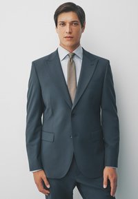 Dunkelblauer Anzug mit Reverskragen, einreihigem Design, kombiniert mit einem hellgestreiften Hemd und einer gemusterten braunen Krawatte.