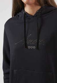 Gros plan d'une personne portant un hoodie noir avec "Amour" brodé et trois petits cœurs en strass en dessous, cordons noirs et poche avant.