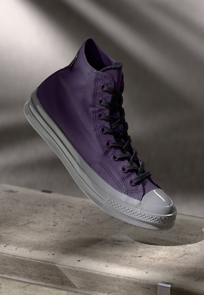 Augstas virsotnes purpura auduma sporta apavi ar melnām saitēm, gumijas pirksta daļu un caurspīdīgu zoli, ar gludu tekstūru un minimālistisku dizainu.