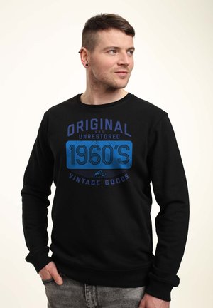 Uomo con felpa nera con testo blu "Original and Unrestored 1960's Vintage Goods", che guarda a destra con la mano nella tasca.