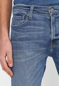 Jeans de mezclilla azul con un acabado desvanecido, diseño de cinco bolsillos y cierre de botón metálico. Incluye costuras visibles y ligeras variaciones en la textura.