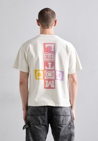 Kortermet kremfarget t-skjorte i bomull med et stort flerfarget grafisk design på ryggen, med fete bokstaver i rosa, lilla og gul.