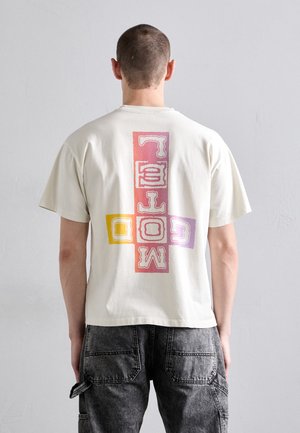 Kortermet kremfarget t-skjorte i bomull med et stort flerfarget grafisk design på ryggen, med fete bokstaver i rosa, lilla og gul.