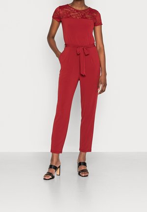 Combinaison rouge avec des manches courtes en dentelle, une taille cintrée accentuée par une ceinture et des jambes fuselées. Portée avec des sandales à talons blocs noires.