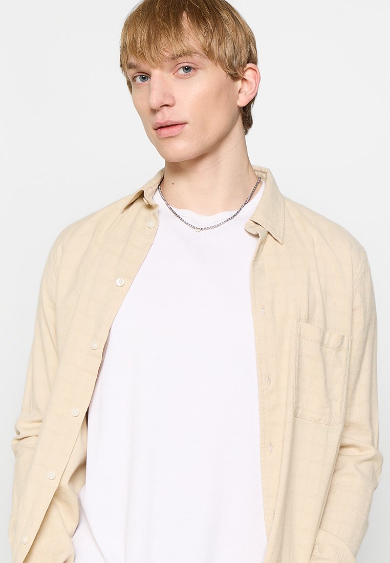 Helle beigen Button-Up-Hemd über einem weißen T-Shirt, mit einer Tasche und subtilem strukturiertem Muster. Das Modell hat kurzes blondes Haar.