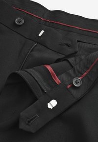 Mustat housut, joissa on sileä kankaan rakenne, vetoketjulla ja napilla varustettu kiinnitys, sekä viininpunaiset sisäkoristeet vyötärön lähellä.