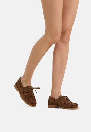 Legs wearing brown suede moccasin shoes with laces, positioned with one foot slightly raised against a plain background.