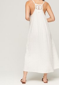 Vestido maxi blanco con un respaldo de ganchillo, que presenta tirantes finos y una tela texturizada. El dobladillo tiene un sutil borde festoneado.