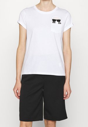 Une personne porte un t-shirt blanc à manches courtes avec des revers roulés et une petite poche ornée d'un visage de dessin animé avec des lunettes de soleil, associé à un short noir à hauteur des genoux.