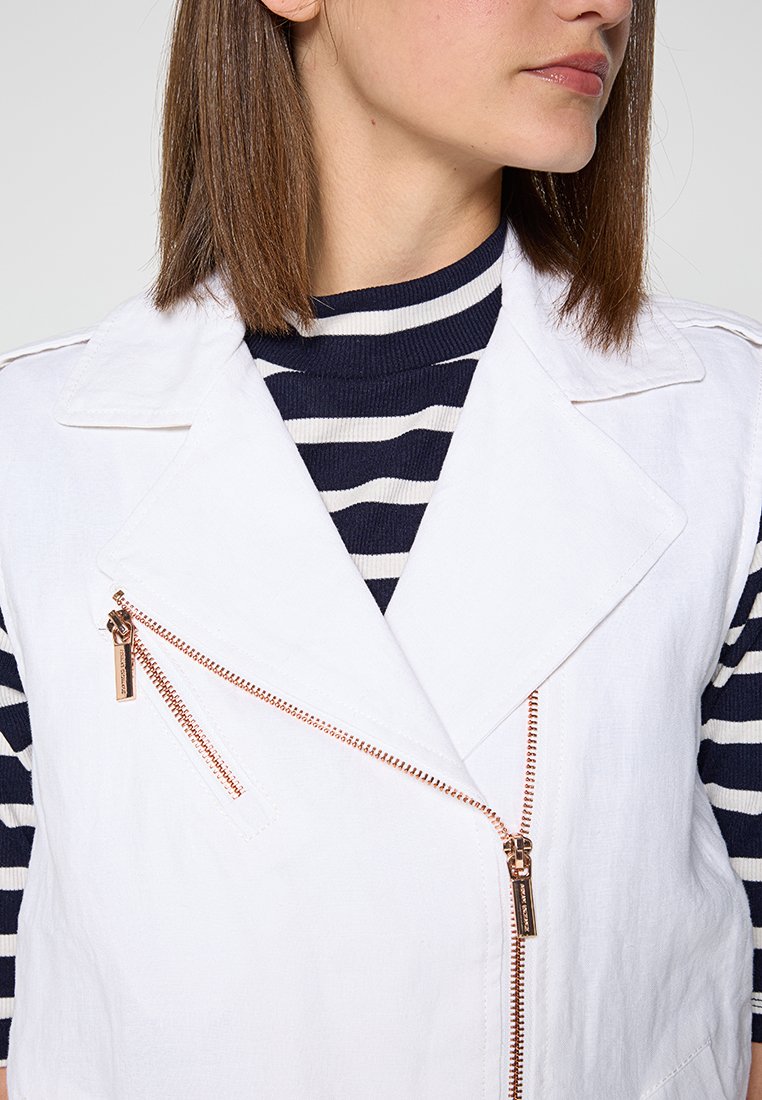 Gilet bianco senza maniche con cerniera diagonale, indossato sopra un dolcevita a righe blu e bianche. Tessuto liscio, design del colletto affilato.