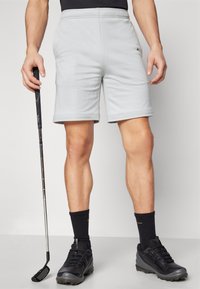 Világosszürke, elasztikus derékpánttal ellátott rövidnadrág, sima textúrával és felgöngyölt szegélyekkel. Fekete sportcipő és kezében tartott golfütő.