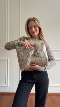 Junge Frau lächelt und hält ein beiges GAP-Shirt in transparenter Plastikverpackung, steht drinnen vor einer weißen Wand mit Verkleidung.