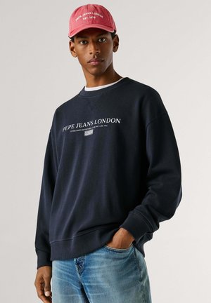 Tumesinine dressipluus valge tekstiga "PEPE JEANS LONDON." Sobitatud roosa nokamütsiga, millel on sarnane tekst. Omadused: lõdva lõikega ja pikkade varrukatega.