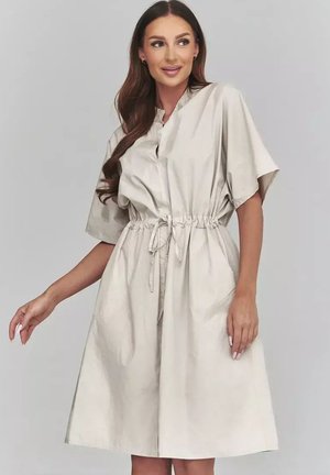 Kobieta o długich brązowych włosach, ubrana w jasno-beżową sukienkę o krótkich rękawach, sięgającą do kolan, z przewiązaną talią, stojąca na jednolitym tle.