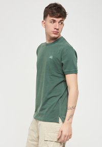 T-shirt verde in cotone con collo rotondo, maniche corte, spacchi laterali e un piccolo dettaglio del logo sul petto. Tessuto morbido, vestibilità casual.