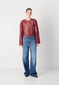 Raudona odinė striukė su dėvėta siluetu, užtrauktukų detalėmis ir diržo juosmeniu, derinama su šviesiai mėlynais plačiais džinsais ir juodais smailiais bateliais.