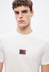 Λευκό βαμβακερό T-shirt με ορθογώνιο μαύρο και κόκκινο μπαλώματα, στο οποίο αναγράφεται το κείμενο "HUGO" κεντραρισμένο στο στήθος. Απλός σχεδιασμός με στρογγυλή λαιμόκοψη.