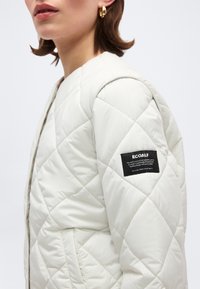 Γυναίκα με λευκό μπουφάν καπιτονέ με κεντημένη ετικέτα της μάρκας ECOALF στη μανσέτα και χρυσούς κρίκους στα αυτιά, σε λευκό φόντο.