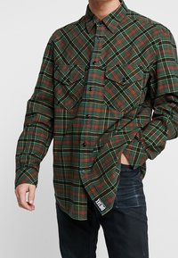 Man med grön, röd och gul rutiga skjorta med knappar och bröstfickor, en hand i fickan på mörka jeans, Diesel-märket synligt.