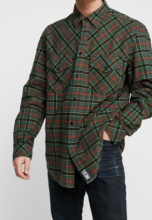 Hombre con una camisa de cuadros en verde, rojo y amarillo, con botones y bolsillos en el pecho, una mano en el bolsillo de unos jeans oscuros, etiqueta de la marca Diesel visible.