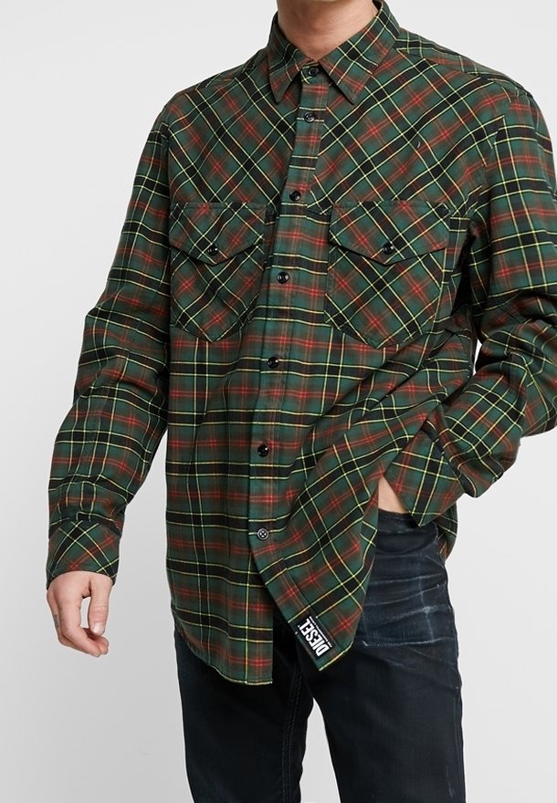 Man med grön, röd och gul rutiga skjorta med knappar och bröstfickor, en hand i fickan på mörka jeans, Diesel-märket synligt.