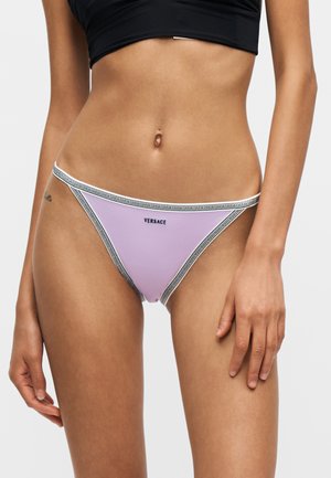 Femme portant un bas de bikini Versace lavande avec un contour à motifs et un crop top noir, debout devant un fond uni.