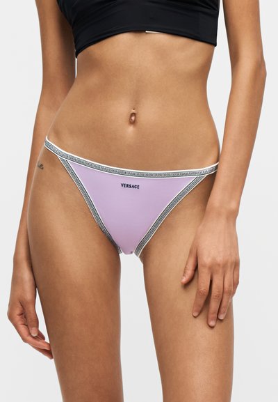 Femme portant un bas de bikini Versace lavande avec un contour à motifs et un crop top noir, debout devant un fond uni.