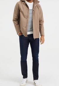 Giubbotto impermeabile color beige con cappuccio, maglione bianco a righe blu navy sotto, jeans in denim blu scuro e sneakers bianche con dettagli neri.
