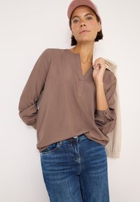 Blusa marrone leggera con scollo a V e dettagli con bottoni, abbinata a jeans blu. La modella tiene una giacca leggera su una spalla.