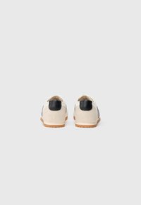 White suede shoes with black leather accents and a gum sole. Features a simplistic design and rounded shape, suitable for casual wear.
