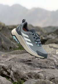 Chaussure de sport grise avec une tige texturée, des rayures blanches, des accents bleu clair et une semelle robuste, marquée "RAIN RDY" sur le côté.