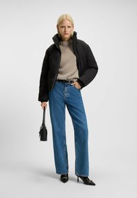 Zwarte gewatteerde pufferjack, beige gebreide trui, high-waisted blauwe jeans en zwarte puntige hakken, met een kleine zwarte handtas.