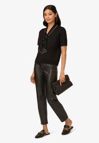 Schwarzes Kurzarmoberteil mit einem Bindungsdetail und weißen Abschlüssen, kombiniert mit schwarzen Kunstlederhosen und schwarzen Slippern, während eine schwarze Clutch gehalten wird.