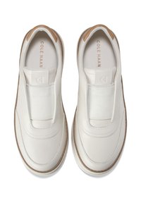 Valkoiset nahkakengät ilman nauhoja, joissa on joustovenyjä päällä ja ruskeat yksityiskohdat kantapäässä. Sisällä merkki "Cole Haan".