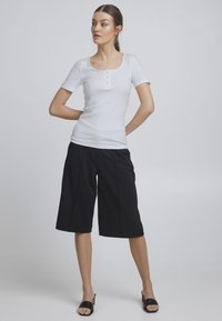 Biela rebrovaná tričko s krátkymi rukávmi a okrúhlym výstrihom, skombinované s čiernymi culottes do kolien a čiernymi šľapkami na podpätku.