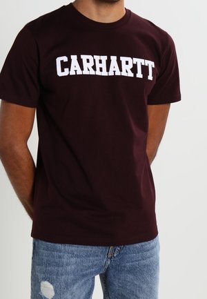 Mann trägt ein dunkelburgunderfarbenes Carhartt-T-Shirt und zerrissene blaue Jeans, steht mit den Händen hinter dem Rücken vor einem schlichten Hintergrund.