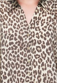 Gros plan d'une chemise beige avec un motif léopard marron foncé et cinq petits boutons ronds dorés au centre devant.
