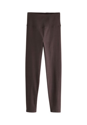 Gros plan d'une personne portant des leggings moulants marron foncé et un haut ample de couleur crème, la main reposant sur la hanche.