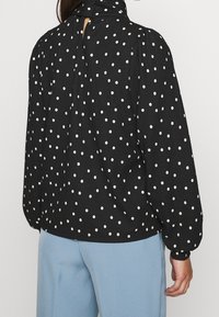 Blusa nera a pois con maniche lunghe, tessuto strutturato, chiusura con bottoni sul retro del collo e dettagli arricciati sulle spalle.