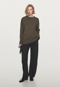 Olivově zelený oversized svetr s žebrovaným výstřihem, kombinovaný s černými kalhotami s širokými nohavicemi a černými botami; model držící černou kabelku.