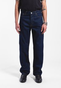 Dark blue denim jeans with a straight fit, featuring orange stitching, a button fly, and front pockets. Worn with black shoes.