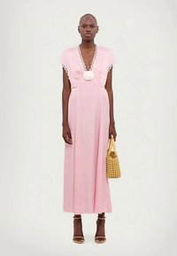 Žena v ružových maxi šatách bez rukávov s bielou čipkovou obrubou, drží tkanú kabelku, na nohách má béžové sandále na vysokom opätku a stojí.