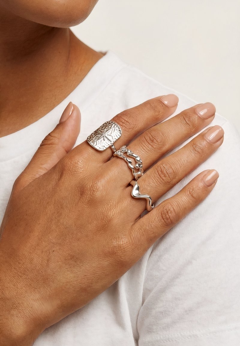 Main avec trois bagues en argent présentant différents motifs organiques et abstraits reposant sur une chemise blanche près de la clavicule.
