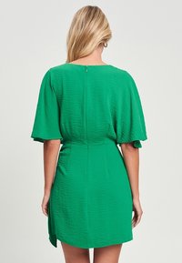 Vihreä mekko, jossa on lyhyet liehuvat hihat, teksturoitu kangas, huomaamaton takana oleva vetoketju ja tiukka vyötärö. Mekossa on minipituinen helma.