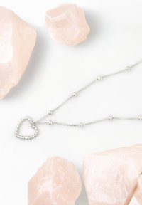 Sølv halskæde med en snoet hjertevedhæng, blandet med runde perler, præsenteret blandt bløde lyserøde krystaller på en hvid baggrund.