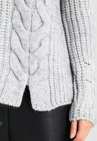 Helles graues Strickpullover mit einem Zopfmuster, geripptem Saum und Ärmeln, Daumenlöchern und lockerer Passform.